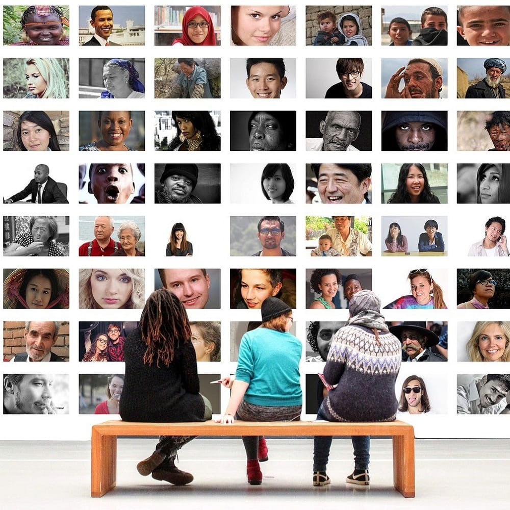 Three people sitting on a bench with their backs to the camera, in front of a wall covered with photographs of people from a diverse range of races and cultures.