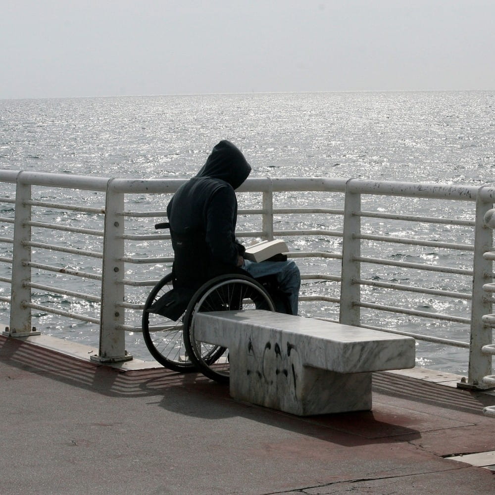 A figure in a black hoodie sitting in a wheelchair with a book open on their lap. Their wheelchair is next to a concrete bench, with a fence in front of them. The sea is visible behind the fence.