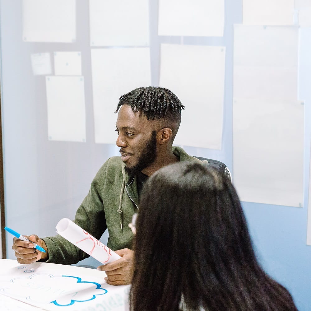A young Black man is sitting at a table with a pen in one hand and some rolled up paper in the other, looking to the left. In the foreground is a person with long dark hair with their back to the camera.