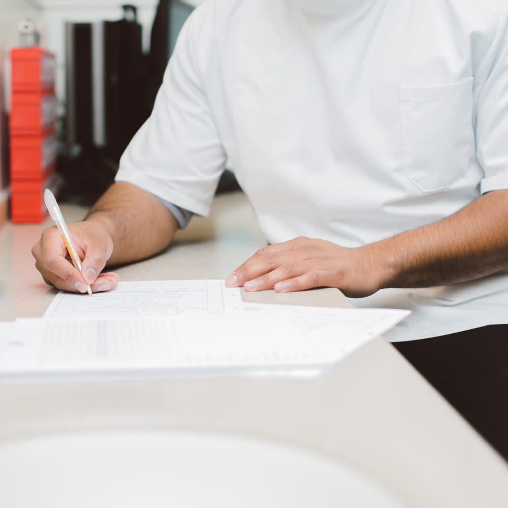 A person, head out of shot, wearing a white tunic with short sleeves. They are turned towards a table next to them. One hand is rested on a piece of paper on the table, and the other is holding a pen, writing something on the paper.