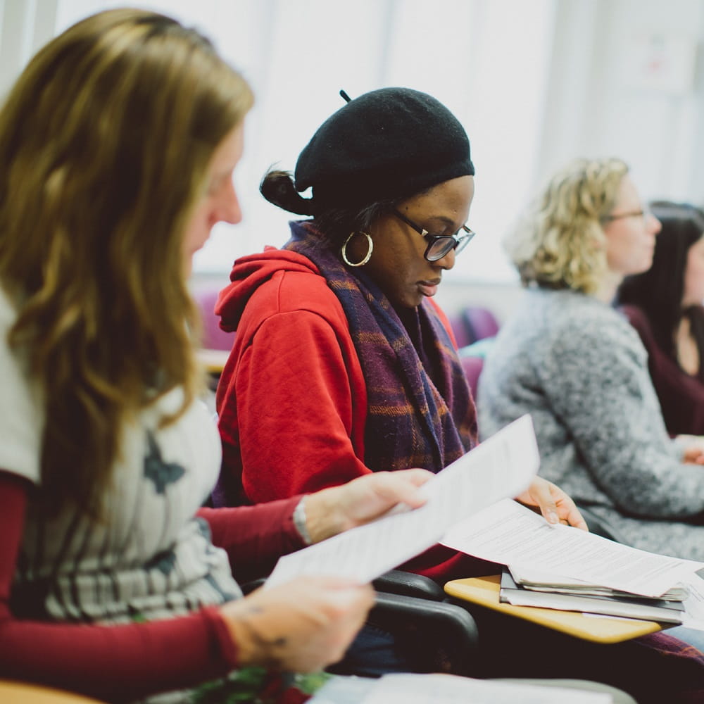 A line of women sitting at seminar chairs with attached desks, with the camera focus on a Black woman in the centre, wearing a beret and looking down at some paper on her desk.
