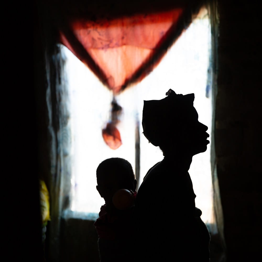A woman wearing a headwrap with an infant on her back is silhouetted by a bright window behind her. A reddish curtain is tied up over the top of the window.