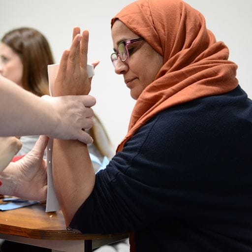 A woman wearing glasses and an orange headscarf watches as someone out of shot uses her arm to demonstrate how to add a splint.
