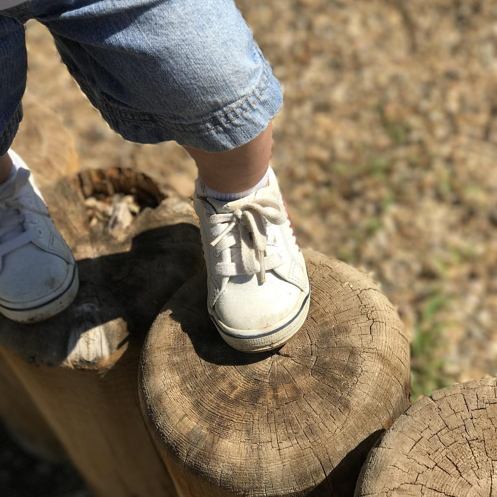 A toddler, mostly out of shot, wearing white shoes and blue shorts, stepping from one wooden step up to another.