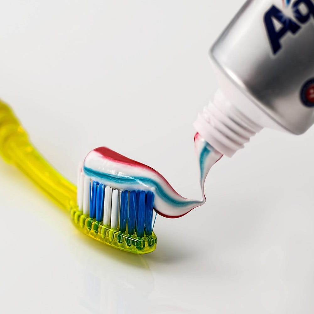 A yellow toothbrush with blue and white bristles, with some blue, red and white toothpaste on the bristles and part of a toothpaste tube on the top right.