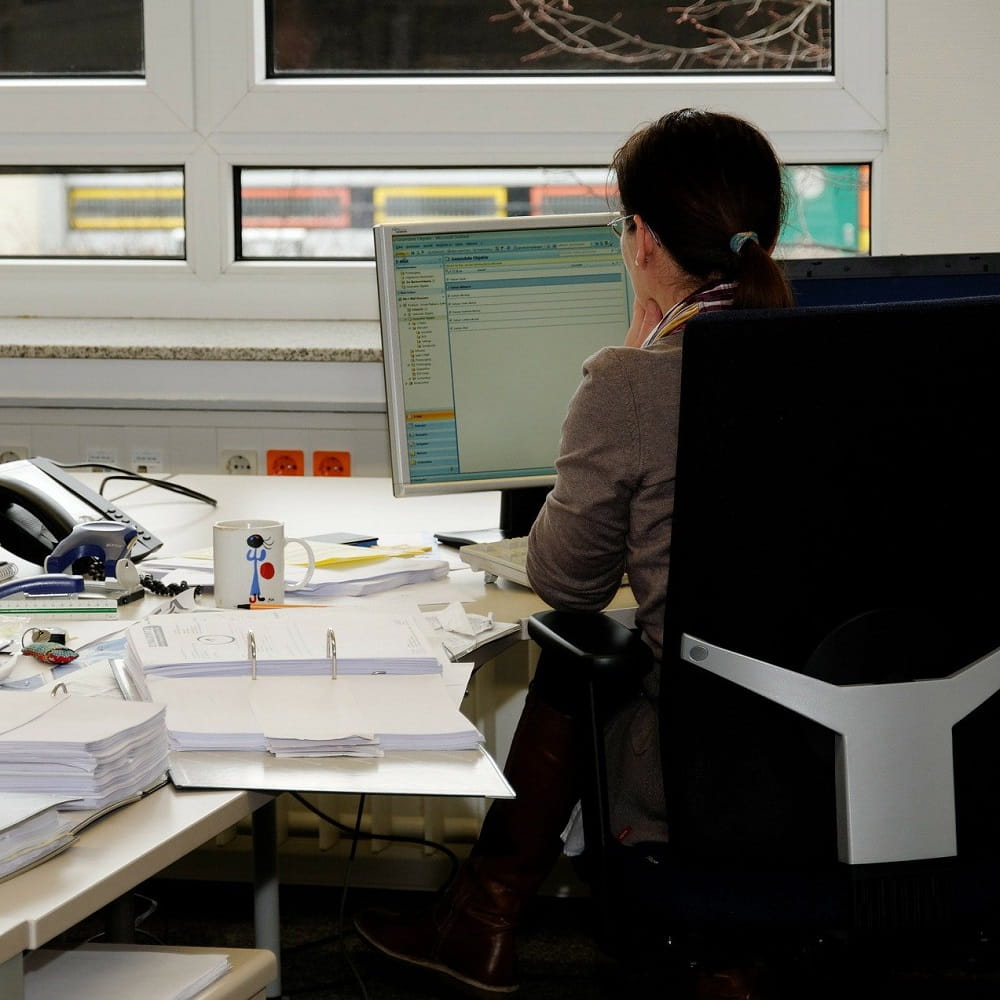 A woman with a ponytail sitting in an office chair at a desk, looking at a computer screen. Her desk is covered in papers and a folder is open next to her.
