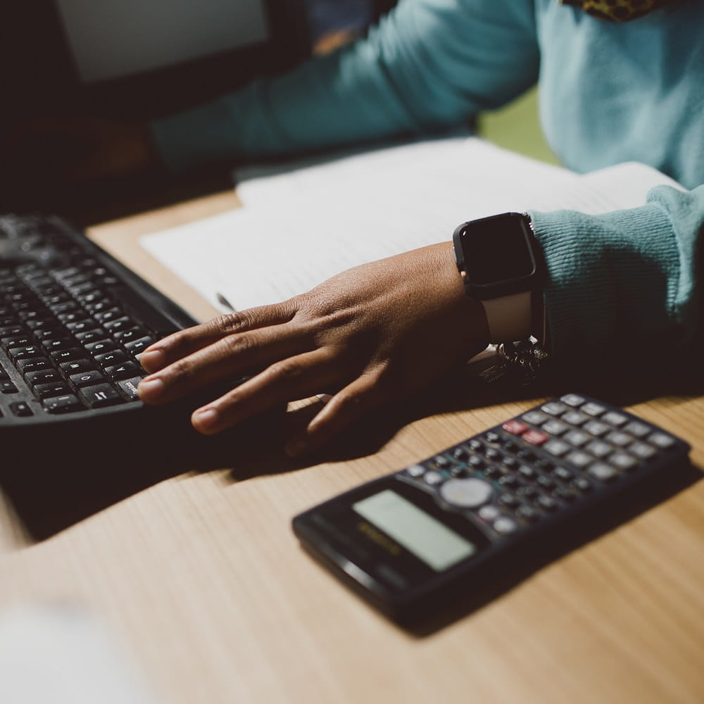 A calculator on a desk next to a hand working at a computer keyboard