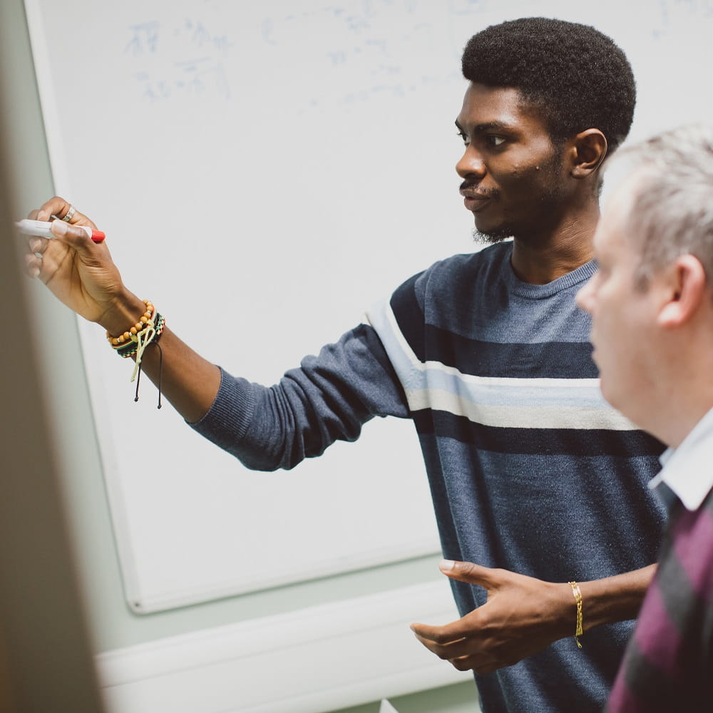 A young Black man holding a board marker is gesturing at the board in front of him to an older white man standing in the foreground.
