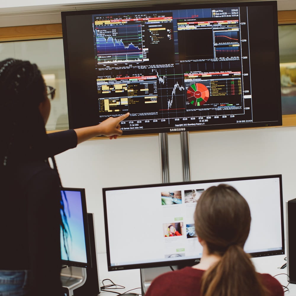 A Black woman is standing on the left, pointing at some data displayed on a large screen on the wall. A white woman is sitting on her right, looking up at the screen she is pointing at.