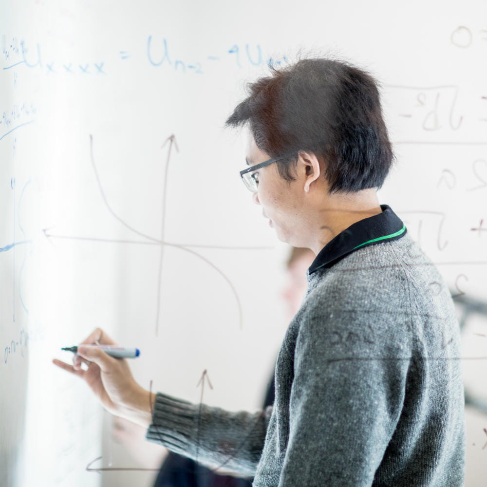 A young man with dark hair and glasses is writing a mathematical formula on a surface, the window through which the photo is taken is also covered in formulas.