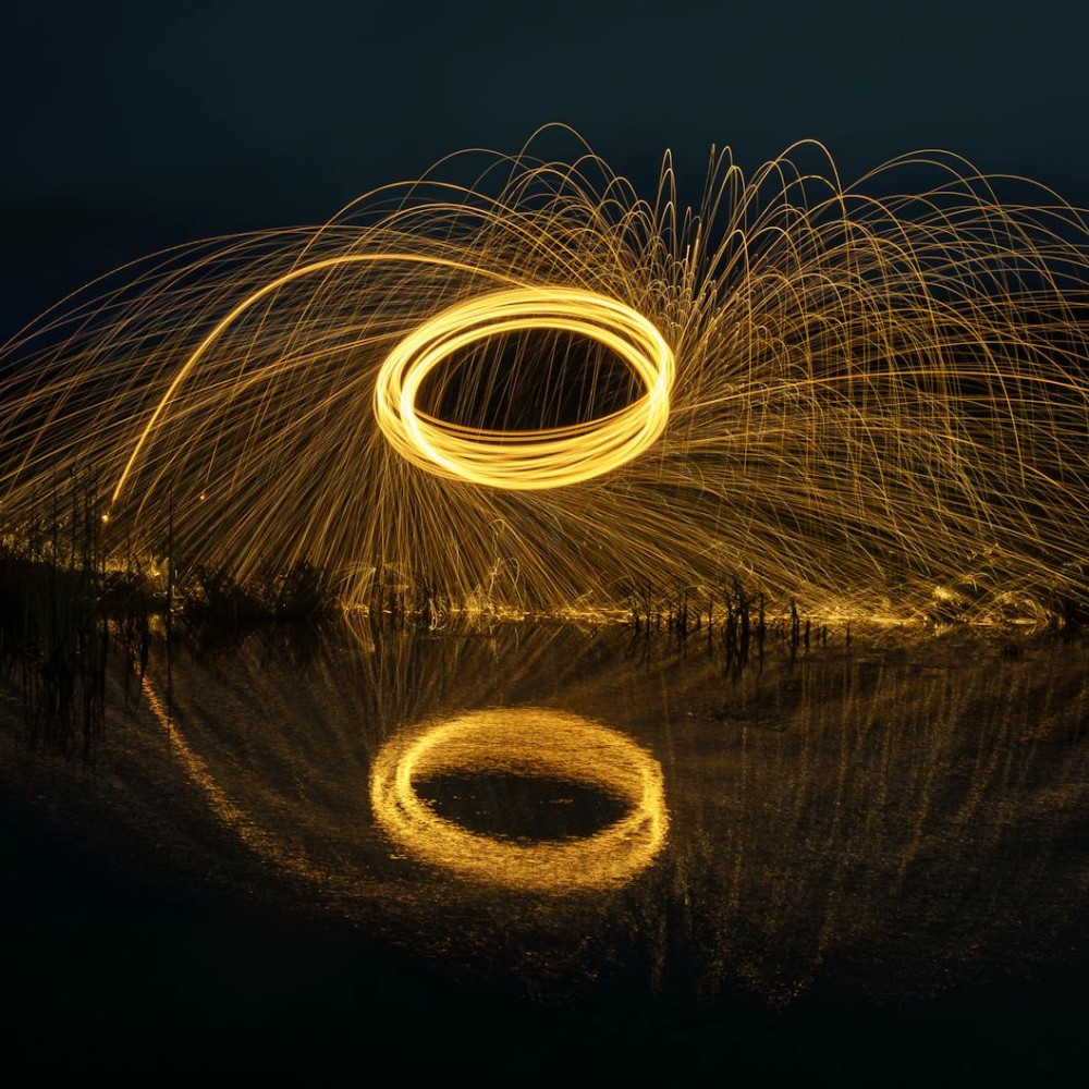 A time-lapsed photo taken at night, showing large yellow circles and sparks created by lit metal wool being spun in a circle.