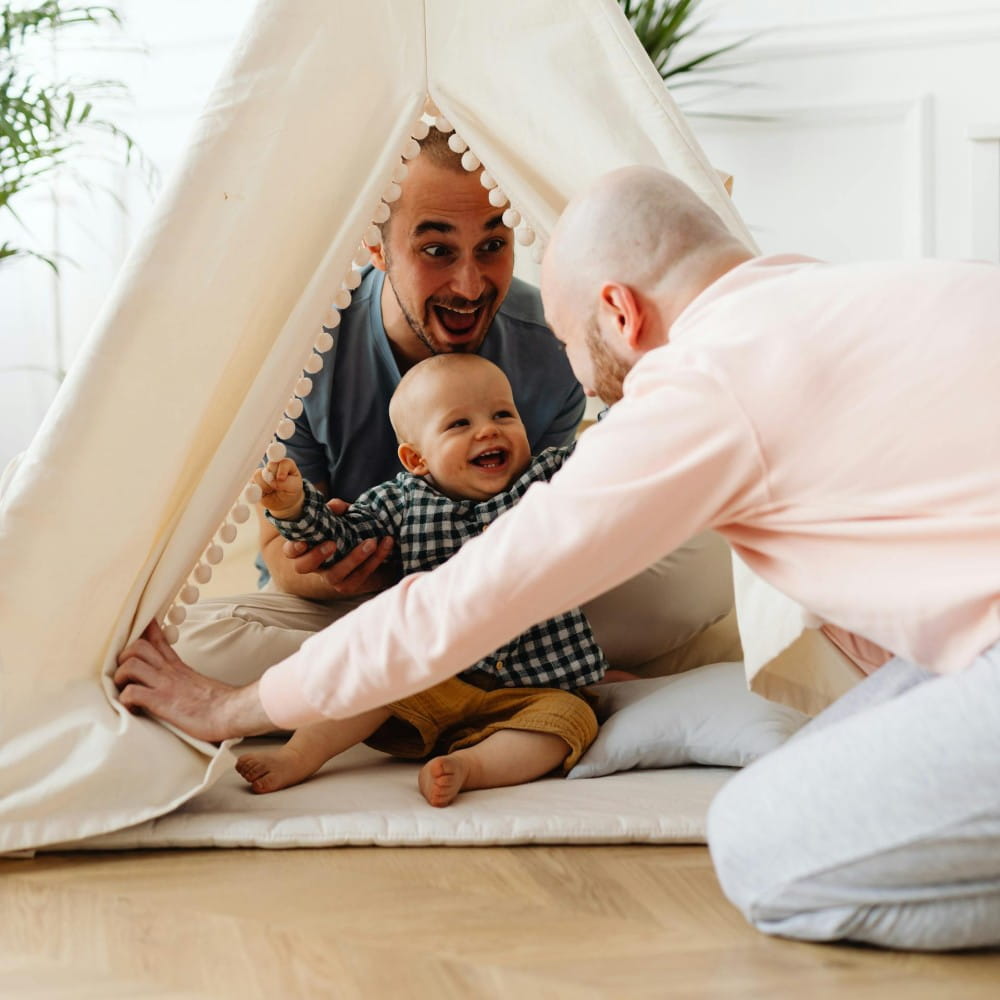 Two Dads, one sitting in a small tent with a baby and the other kneeling in front, playing a game. The baby has a big smile and is holding their arms out to one of the Dads.