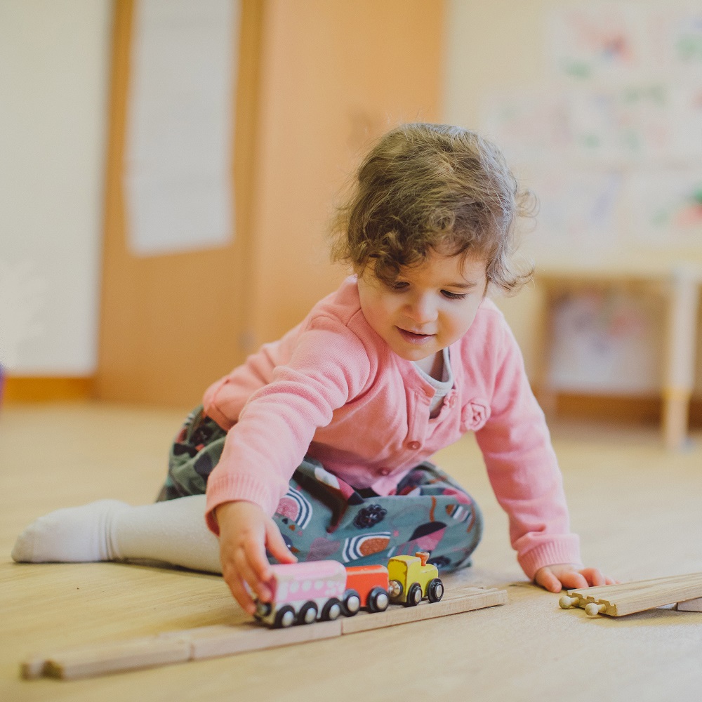 A toddler in a dress and a pink cardigan is sitting on a floor, playing with a little wooden train set.