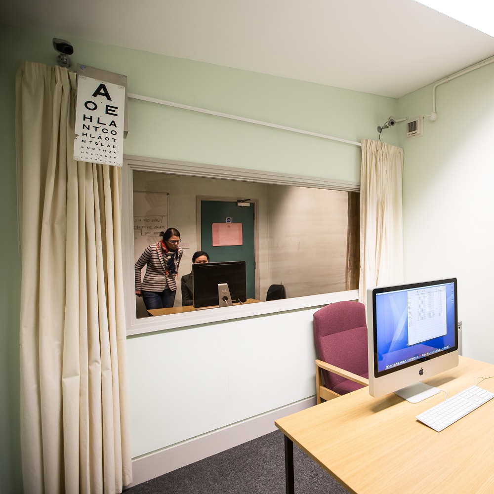 A room with a table and laptop on the right. Towards the back of a room is a mirror window with two people reflected in it. There are curtains either side of the window and an eye chart test hanging on the top left.