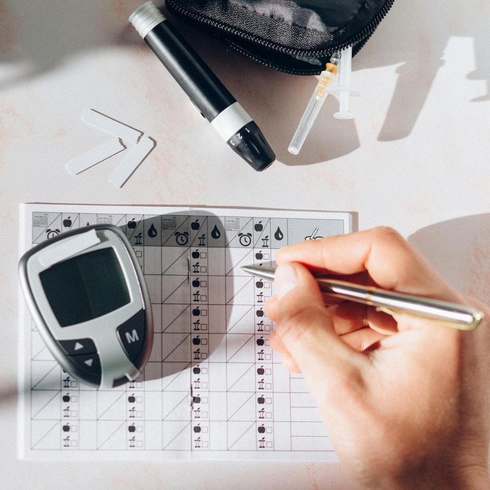 A hand filling out a chart about blood sugars. A small monitor, a lancet and test strips are scattered around.