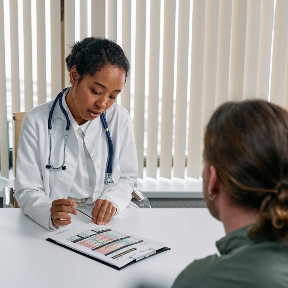 A doctor sitting at a desk looking down at paper on a clipboard, with a patient they are talking to in the foreground.