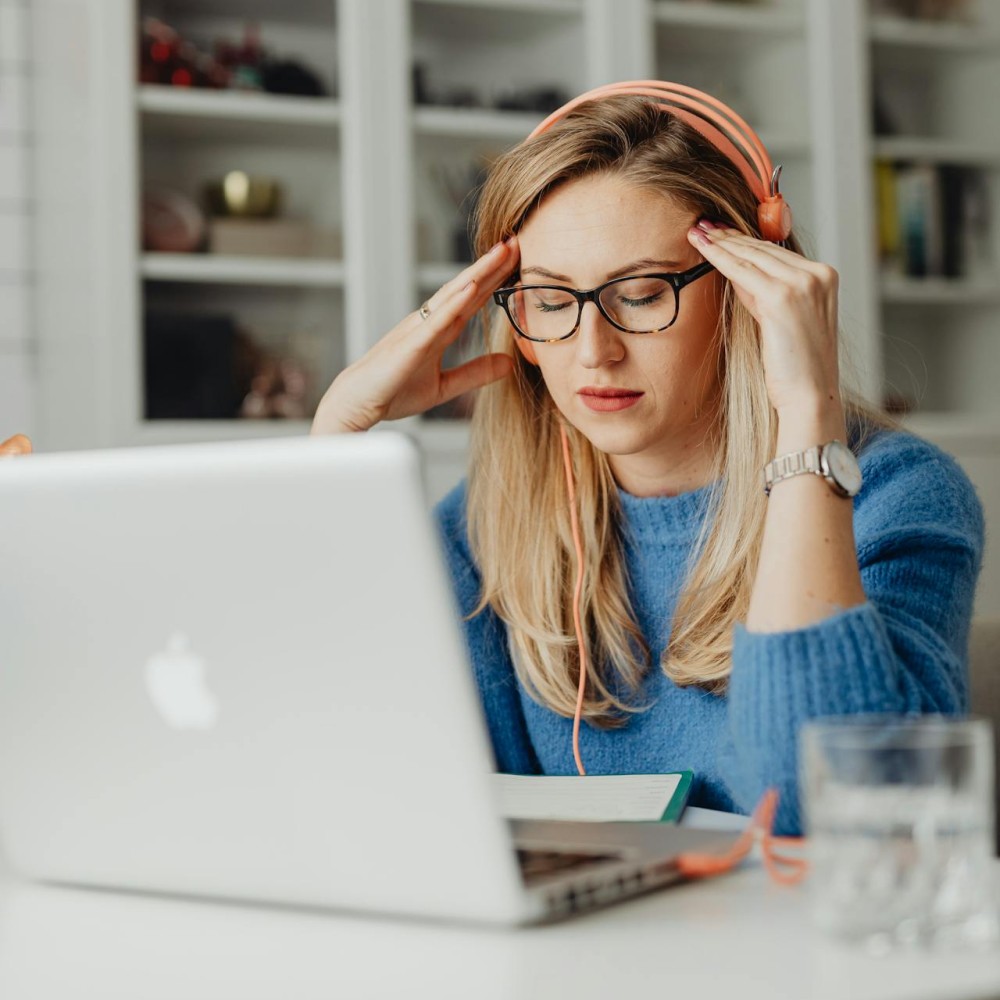 A woman wearing orange headphones sitting in front of a laptop. Her eyes are closed and her fingers are pressed to her temples.