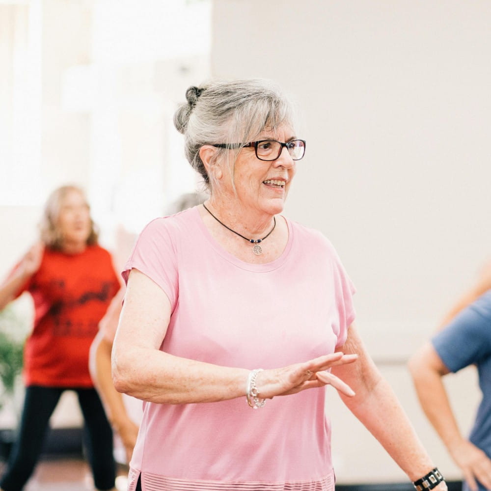 A woman with grey hair wearing a pink t-shirt taking part in an exercise class.