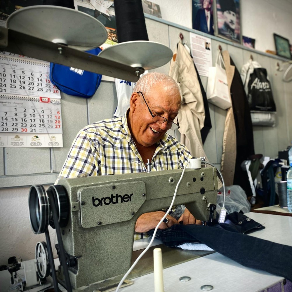 An older man with grey hair sitting in front of a sewing machine, he is looking down at some material in front of him.