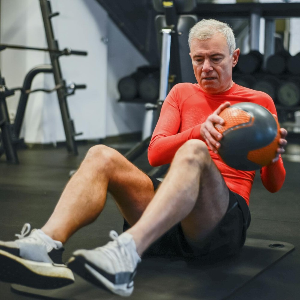 An older man sitting on the floor of a gym, using a ball to exercise.