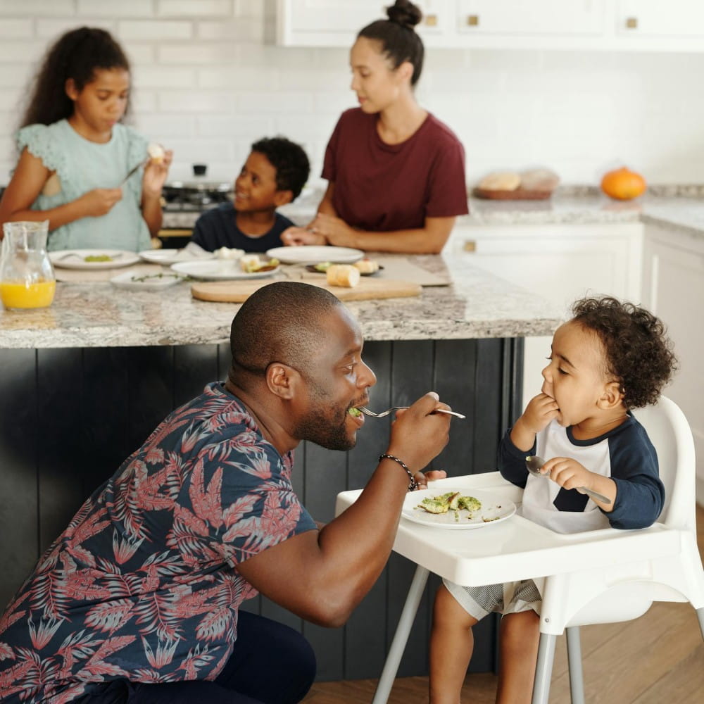 In the foreground a man is crouching in front of a toddler sitting in a highchair, eating some food off a fork. In the background a woman and two more children are standing at a kitchen counter, which has plates of food on it.