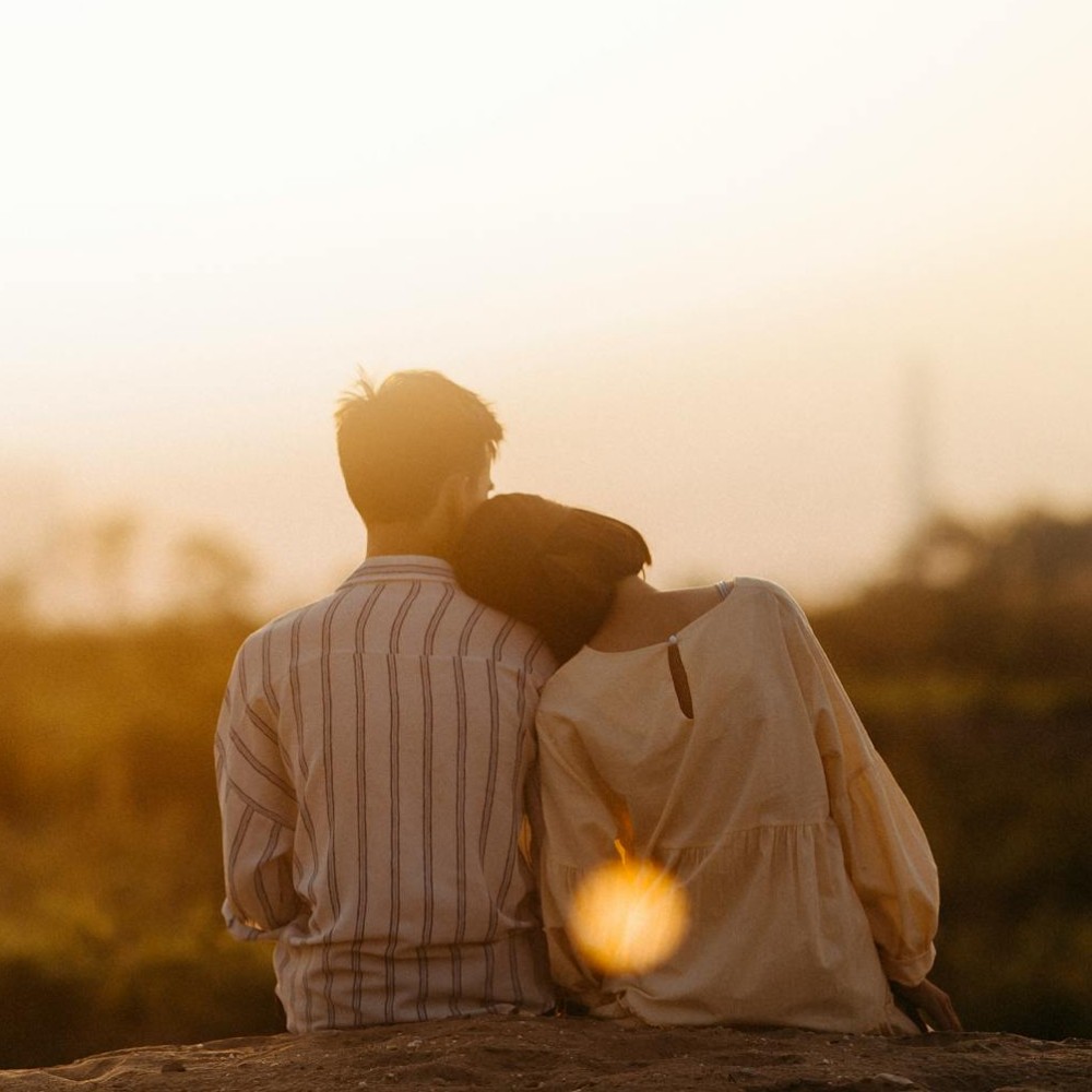 A couple sitting with their backs to the camera. The person on the right is resting their head on the shoulder of the person on the left.