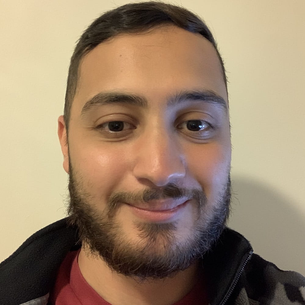 Psychology graduate Hussein Mehmet, with a trimmed beard and moustache, a plain wall in the background.