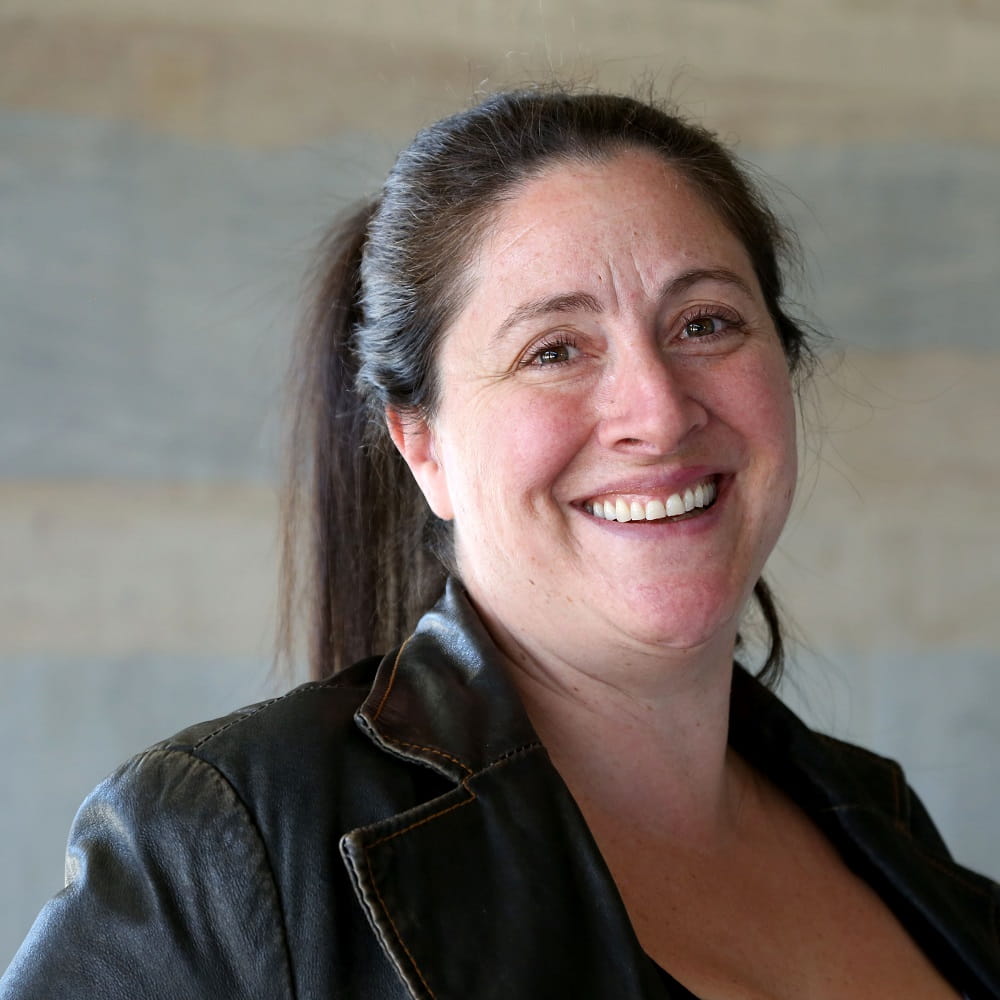 Dr Jordi Asher wearing a black leather jacket and smiling at the camera, with a mottled grey background behind her.