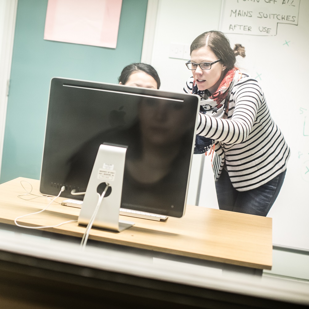 A woman pointing towards a computer screen, with another person's head visible over the top of the computer.