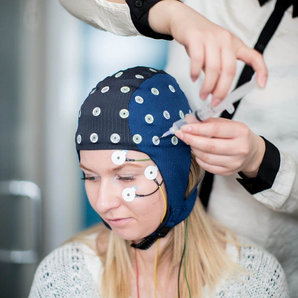 A woman wearing an EEG cap, her head turned away from the camera while another person adjusts the cap.