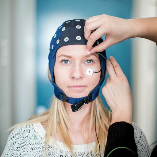 A woman wearing an EEG cap is looking directly at the camera, while a second person mostly out of shot on the right attaches a sensor to her face.