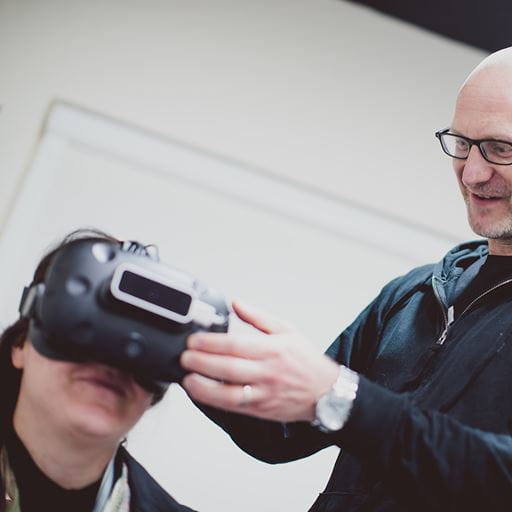 Professor Paul Hibbard stands on the right of the shot, his hands reaching out to adjust a VR headset that a person to the left of the photo is wearing.