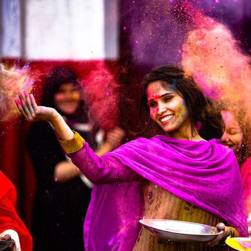 A woman wearing a purple shawl throwing up coloured powder at a Holi festival.