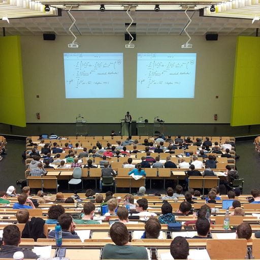 Taken at the back of a lecture hall, with rows of students in seats and maths formulae visible on the projector screen in the background.
