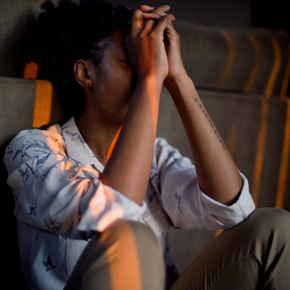 A person sitting with their elbows on their knees, their hands clasped and covering their face.