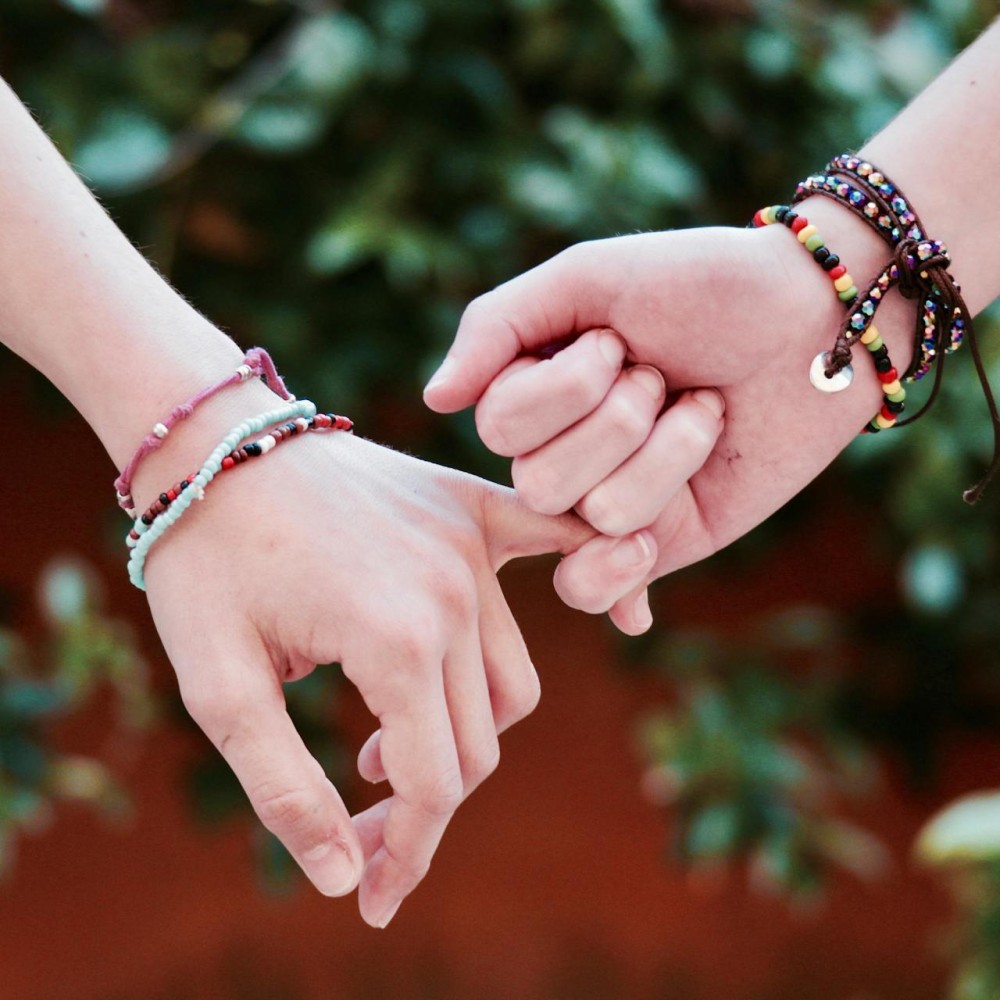 Two hands, one with their pinky finger wrapped around the other. They have beaded bracelets on their wrists.
