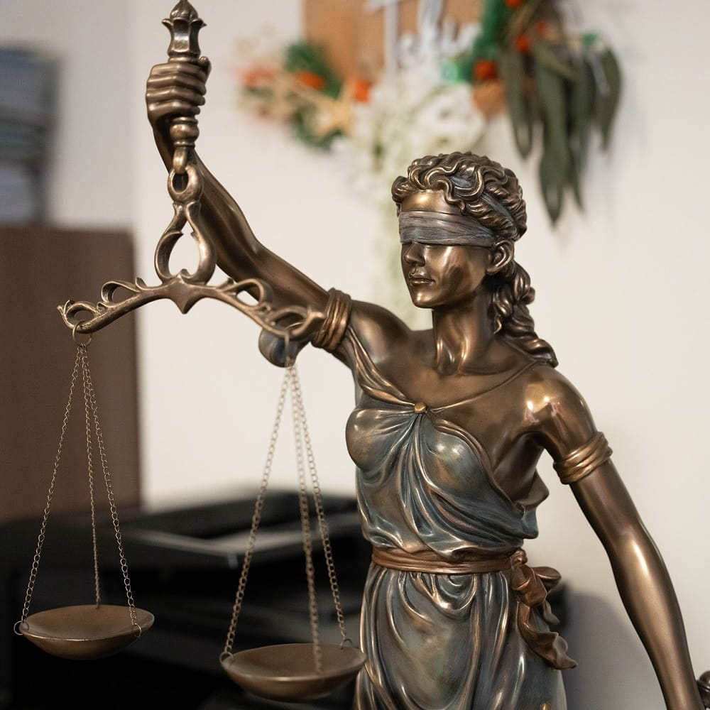 A small metal statue of the figure of "Justice", blindfolded and holding up a pair of scales, with a wall and some artwork visible in the background.