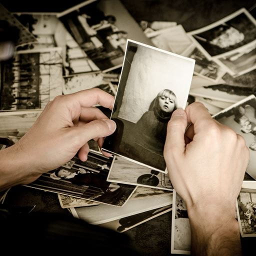 Two hands holding a black and white photo of a child, a messy pile of more black and white photos in the background.