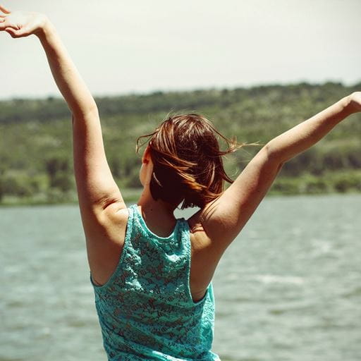 A woman in a green top with her back to the camera, arms raised towards the sky. Water and a hill are in the background.