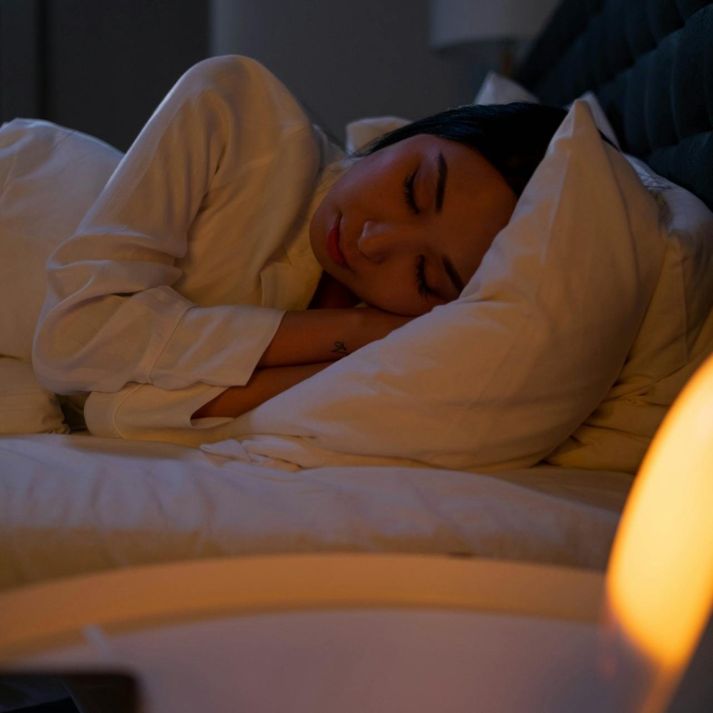 A sleeping woman lying on her side in bed facing the camera. A lamp is in the foreground.