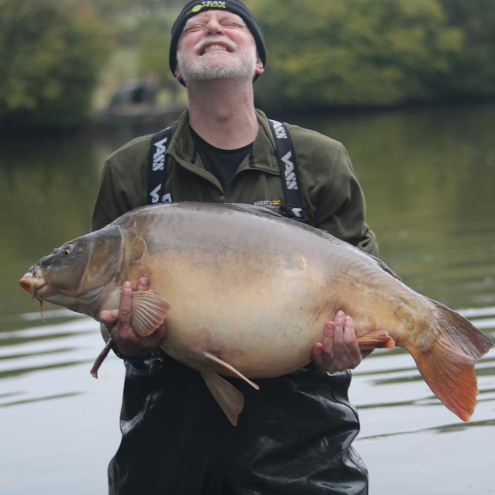 A man standing in water wearing waders and a green shirt is holding a very large fish. His head is tilted towards the sky and he is smiling with his eyes closed.
