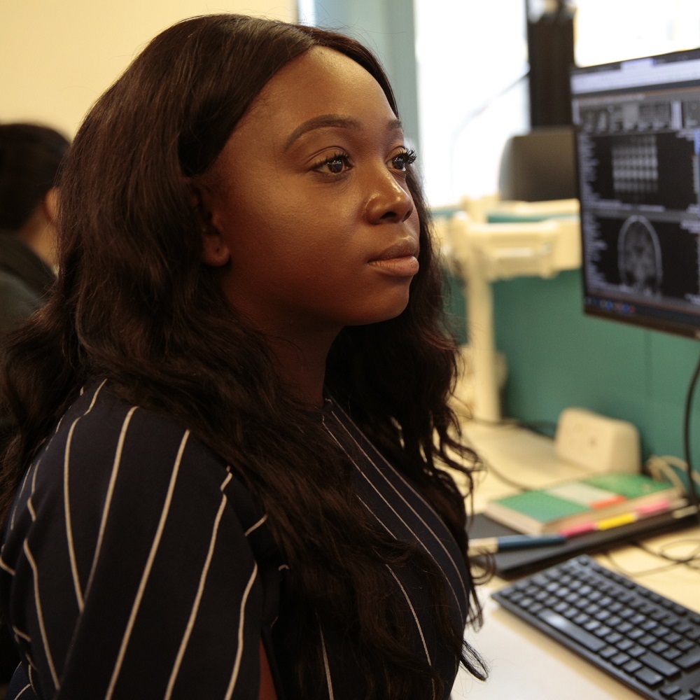 Psychology graduate Sammy Shittu, a young Black woman wearing a black and white shirt, looking at a computer screen out of shot on the right with another computer screen and keyboard visible in the background.