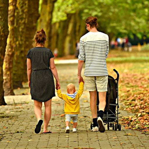 Two people walking down a tree-lined path, holding the hands of a toddler in a yellow coat who is walking between them. The person on the right is pushing a pushchair with their other hand.