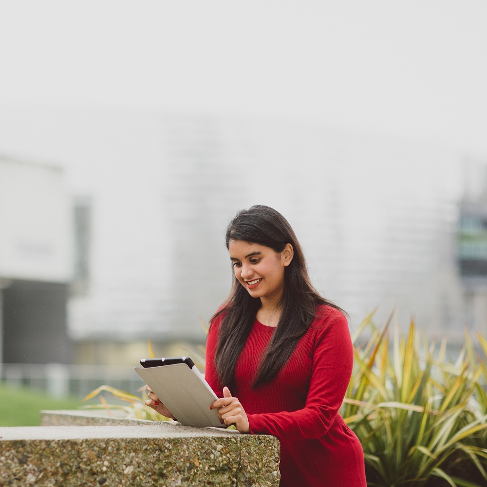 A woman in a red top standing outside looking at an iPad she's holding.