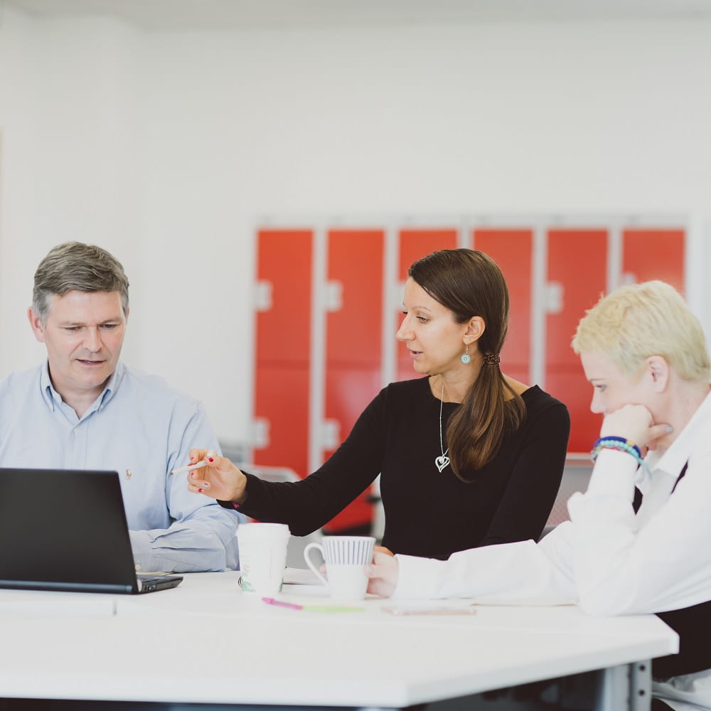 Three people sitting at a desk having a meeting, the person in the middle is pointing at a laptop in front of them. A set of red lockers are visible in the background.