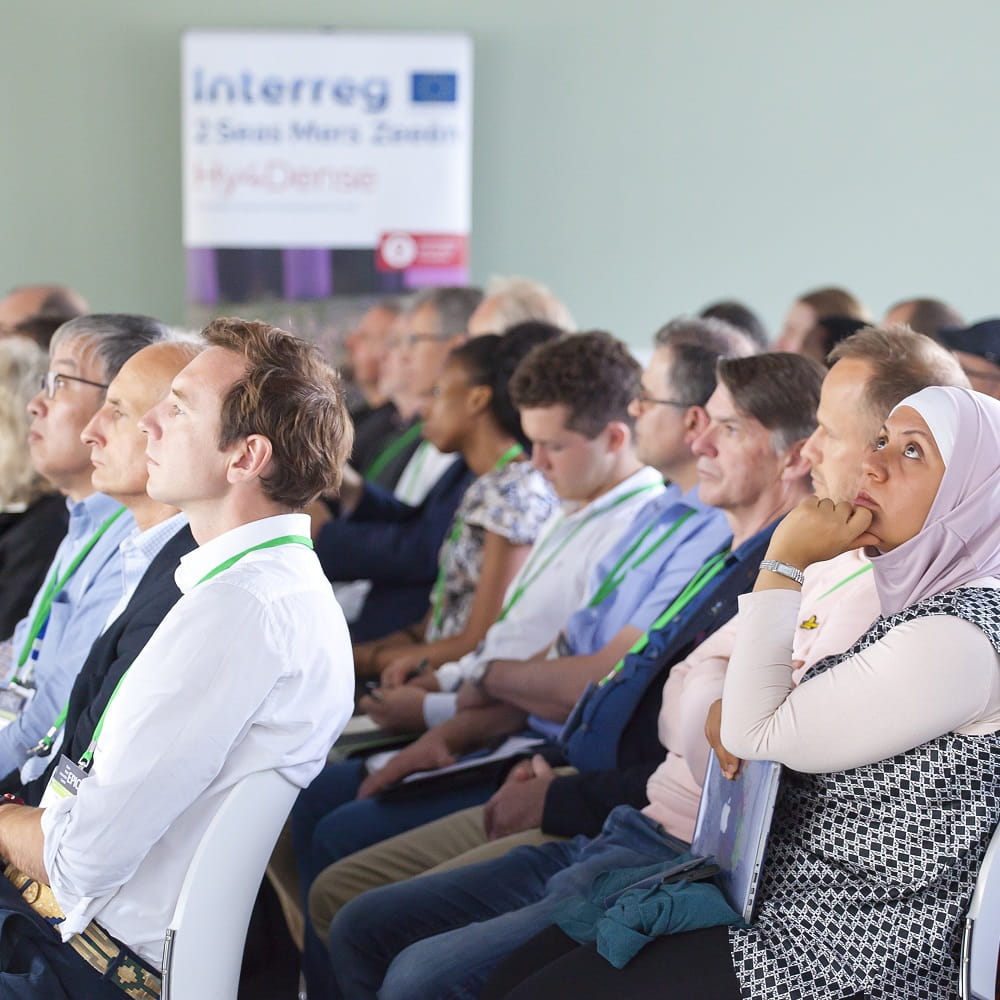 Three rows of people sitting on white chairs. On the right a woman wearing a pink headscarf is looking diagonally up towards something out of shot on the left hand side.