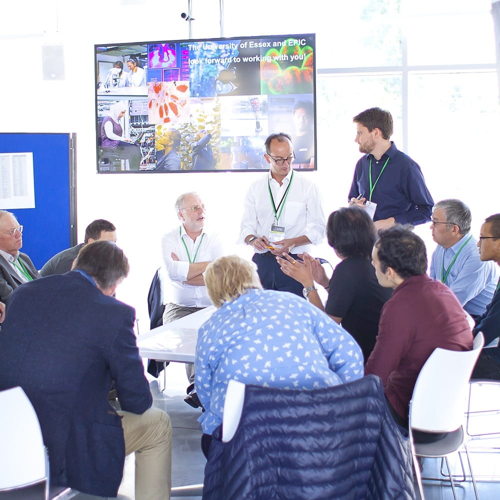 A group of people sitting and standing around a large table, with a TV screen in the background towards the ceiling showing a range of scientific images.
