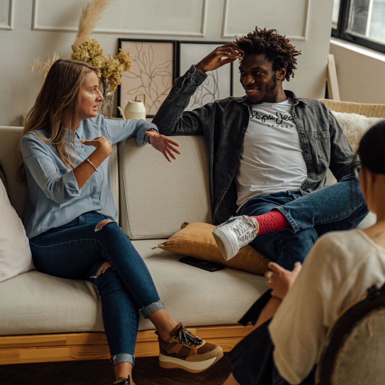Two people sitting on a sofa and talking. A third person with their back to the camera is slightly out of frame on the right.