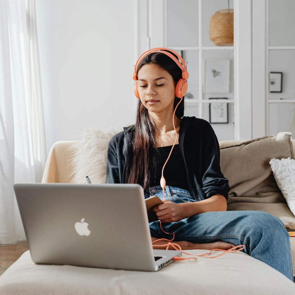 A teenage girl with headphones on is sitting on a sofa looking at a laptop in front of her.