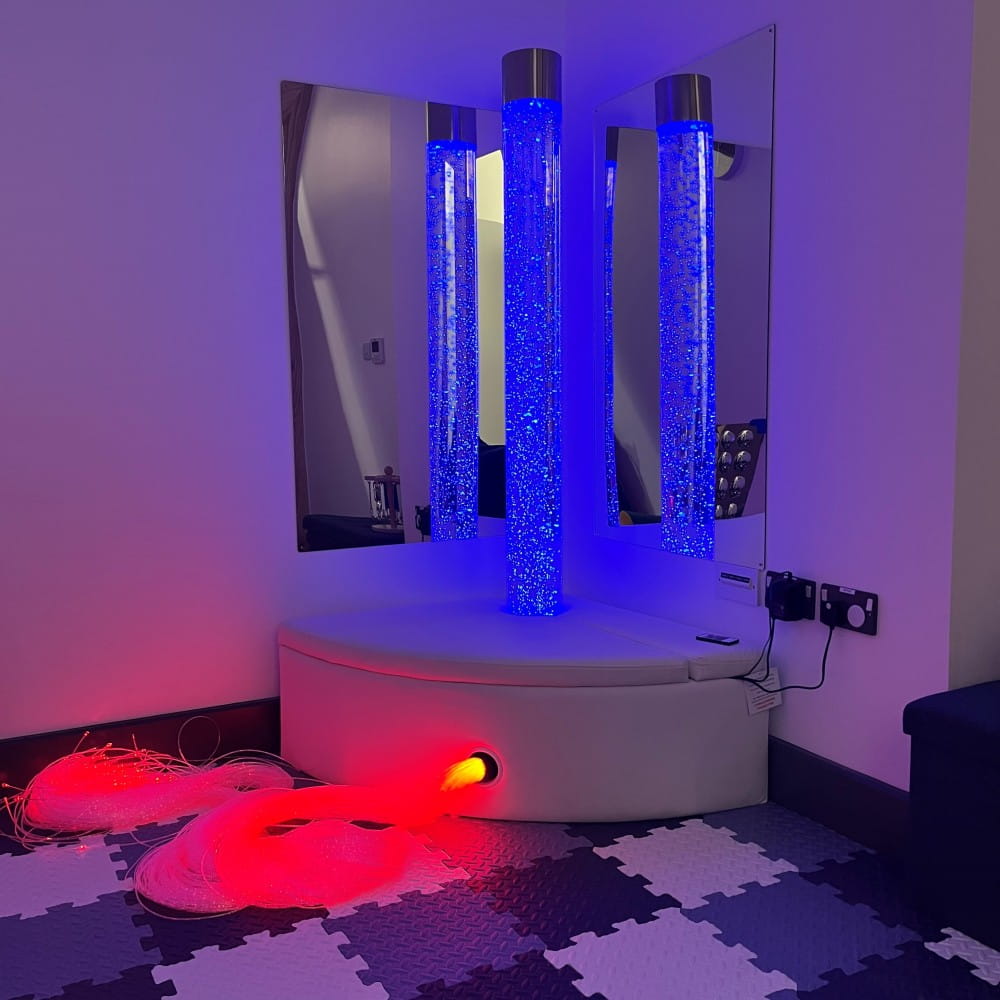 A corner of a sensory room with three bubble tubes lit up in blue light, with mirrors on the wall behind. Some fibre option cable is on the floor, lit up in red.